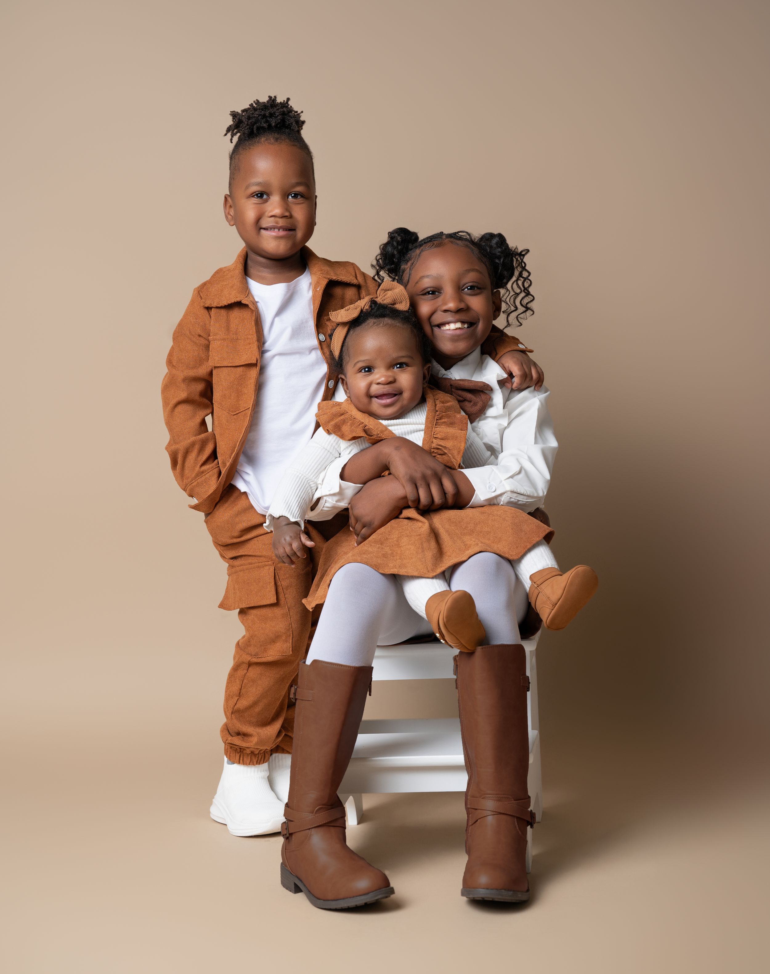 Toddler little kid and big kid posing for their sibling photoshoot in front of a neutral tan backdrop in matching white and brown outfits.