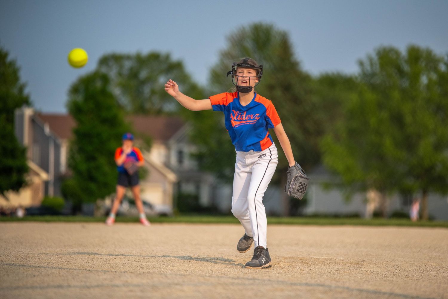 A softball pitcher in a blue and red uniform winds up to throw a yellow ball during a game on a dirt field.