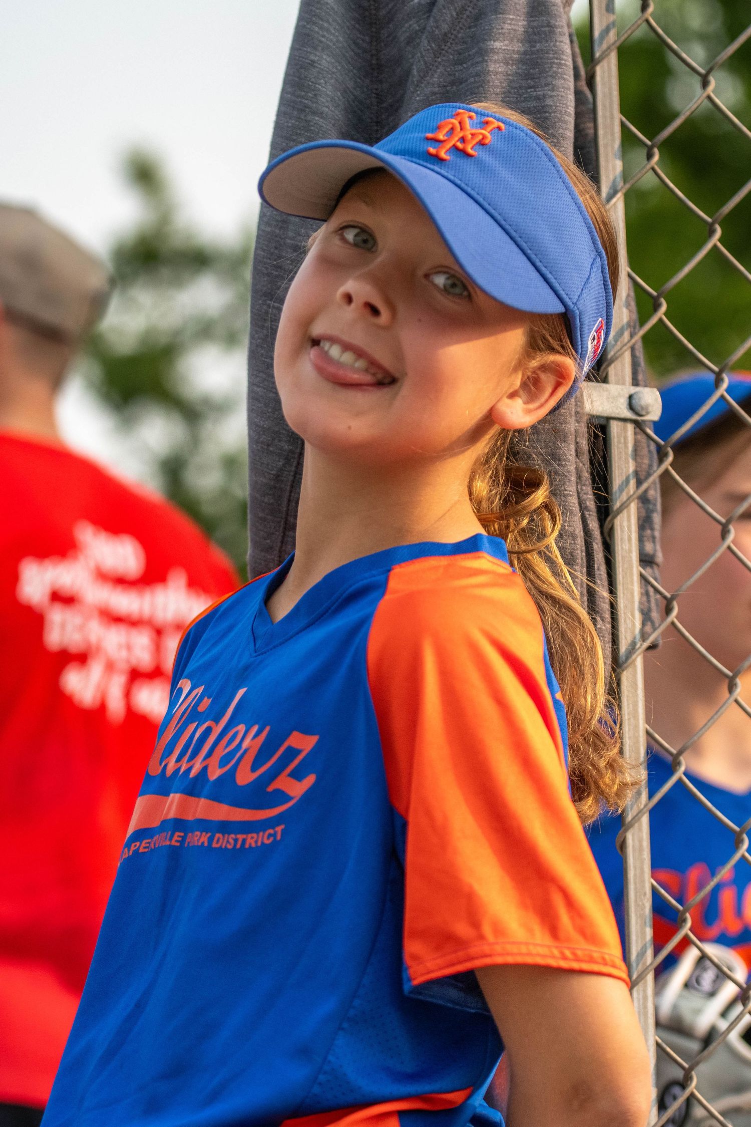 Young baseball player in New York Mets cap and blue and orange uniform smiles while leaning against chain link fence.