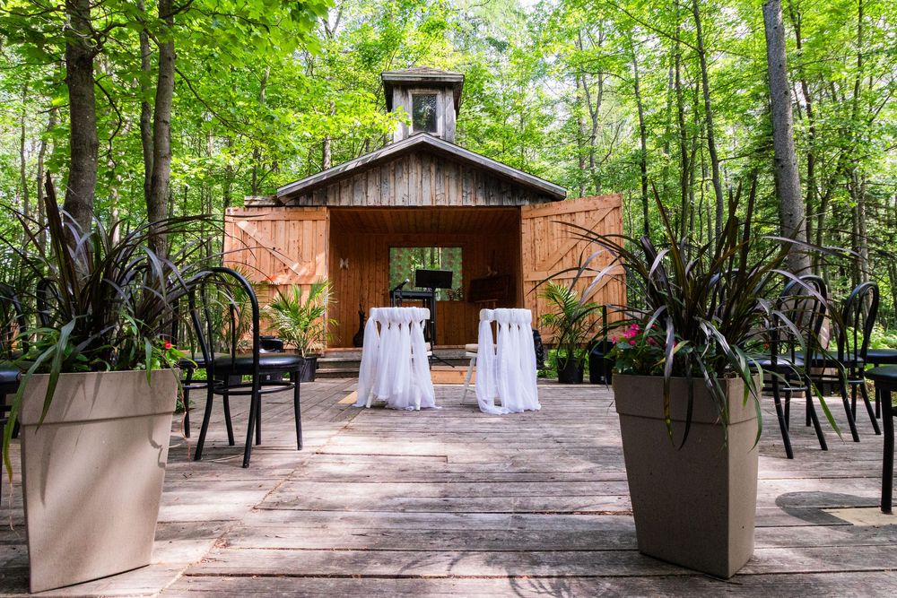 A rustic wooden chapel in a forest setting with black chairs and decorative planters arranged for an outdoor wedding ceremony.