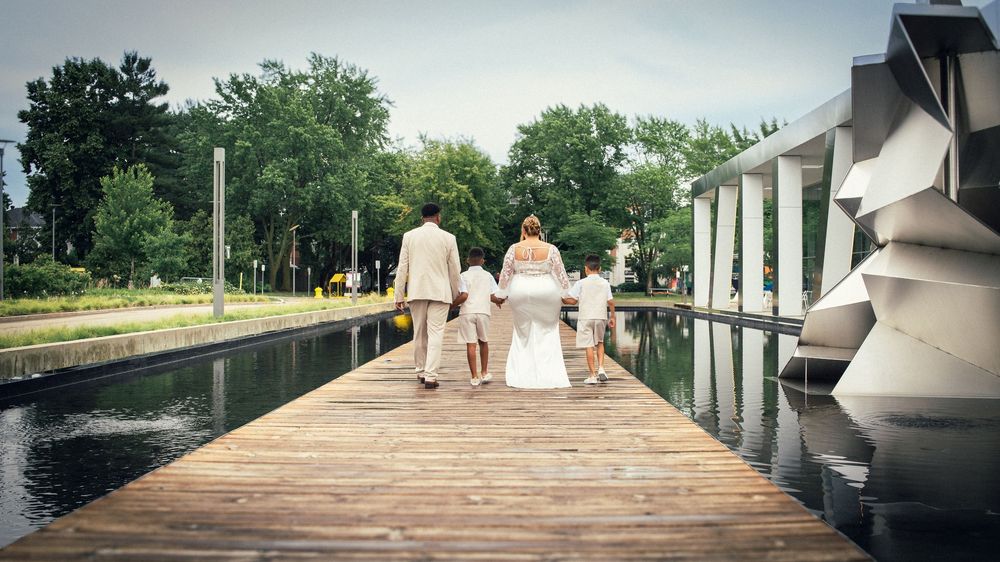 Three people in white outfits walk together on a wooden dock over a tranquil reflecting pool surrounded by trees.