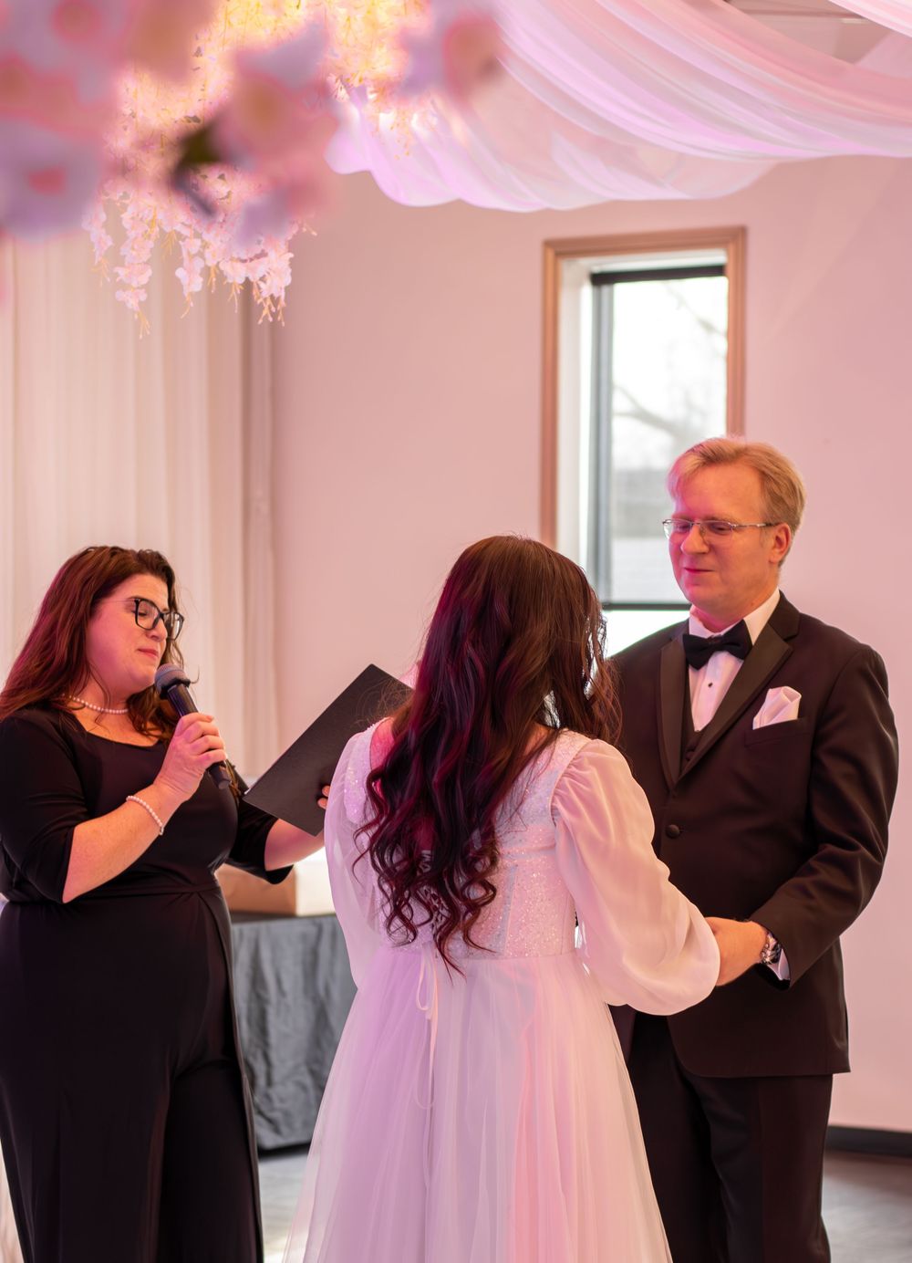Wedding ceremony in progress with officiant speaking to couple under pink lighting and decorations.