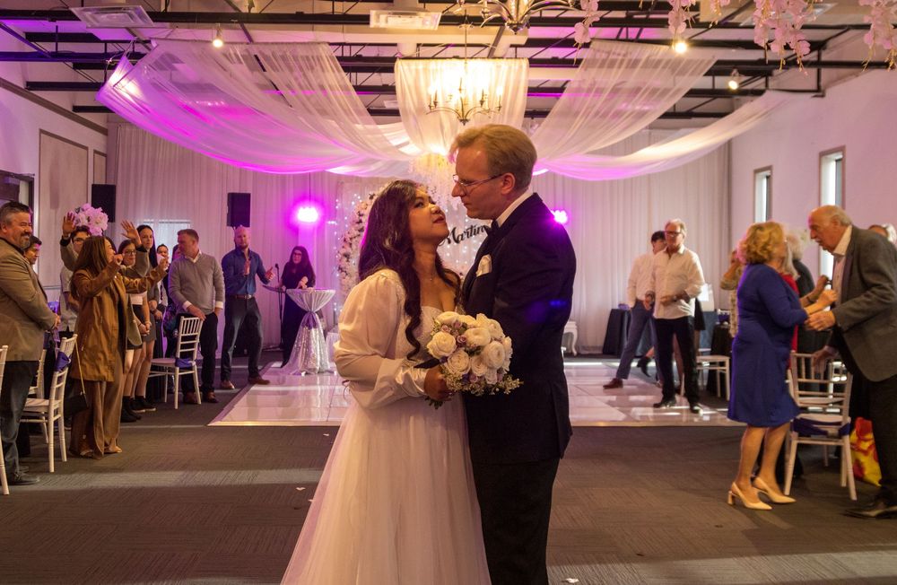 Couple dancing in decorated reception hall with purple lighting and floral arrangements.