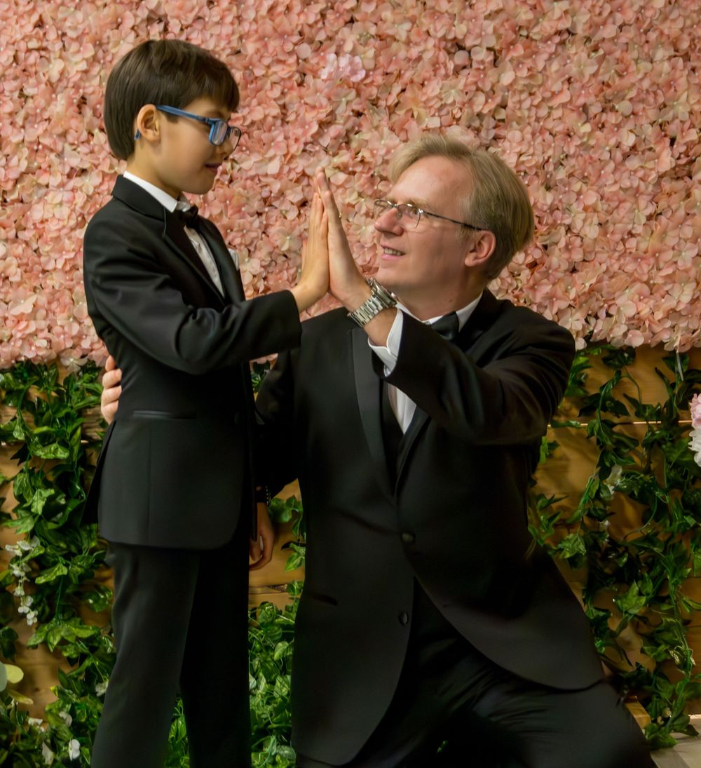 Two people in formal black tuxedos doing celebratory high five against pink floral backdrop.