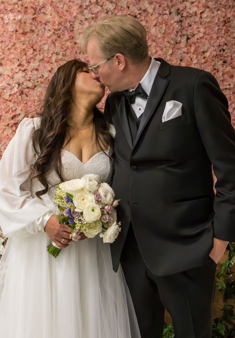 Newlyweds sharing intimate kiss in front of pink floral wall with bride holding white bouquet.