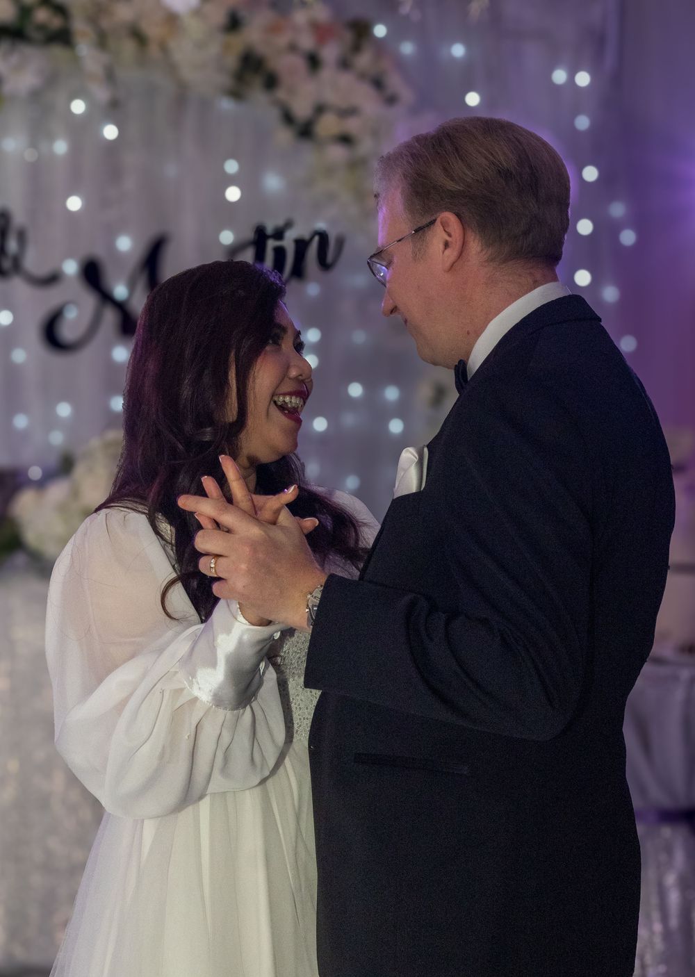 Couple shares joyful dance moment against backdrop of twinkling lights and floral decorations.