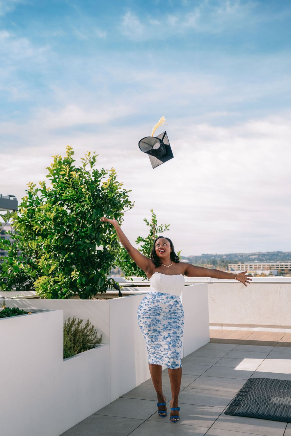 Graduate in white and blue dress celebrates by tossing cap on rooftop terrace.