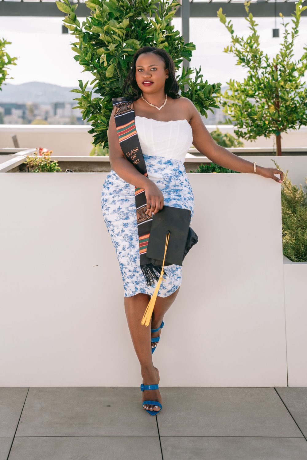 Graduate wearing white and blue dress with sash leans against wall on outdoor terrace.