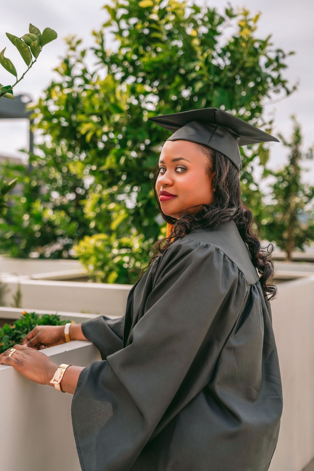 Graduate in black cap and gown poses outdoors against greenery on graduation day.