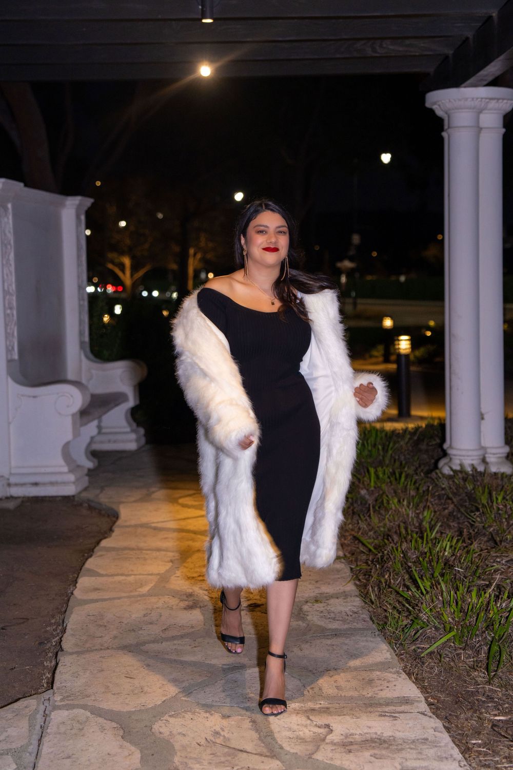 Stylish woman in black dress and white fur coat walks along stone path at night.
