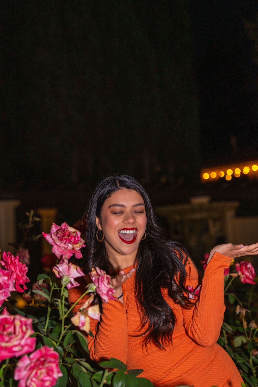 Candid shot of woman in orange dress laughing near pink rose bushes.