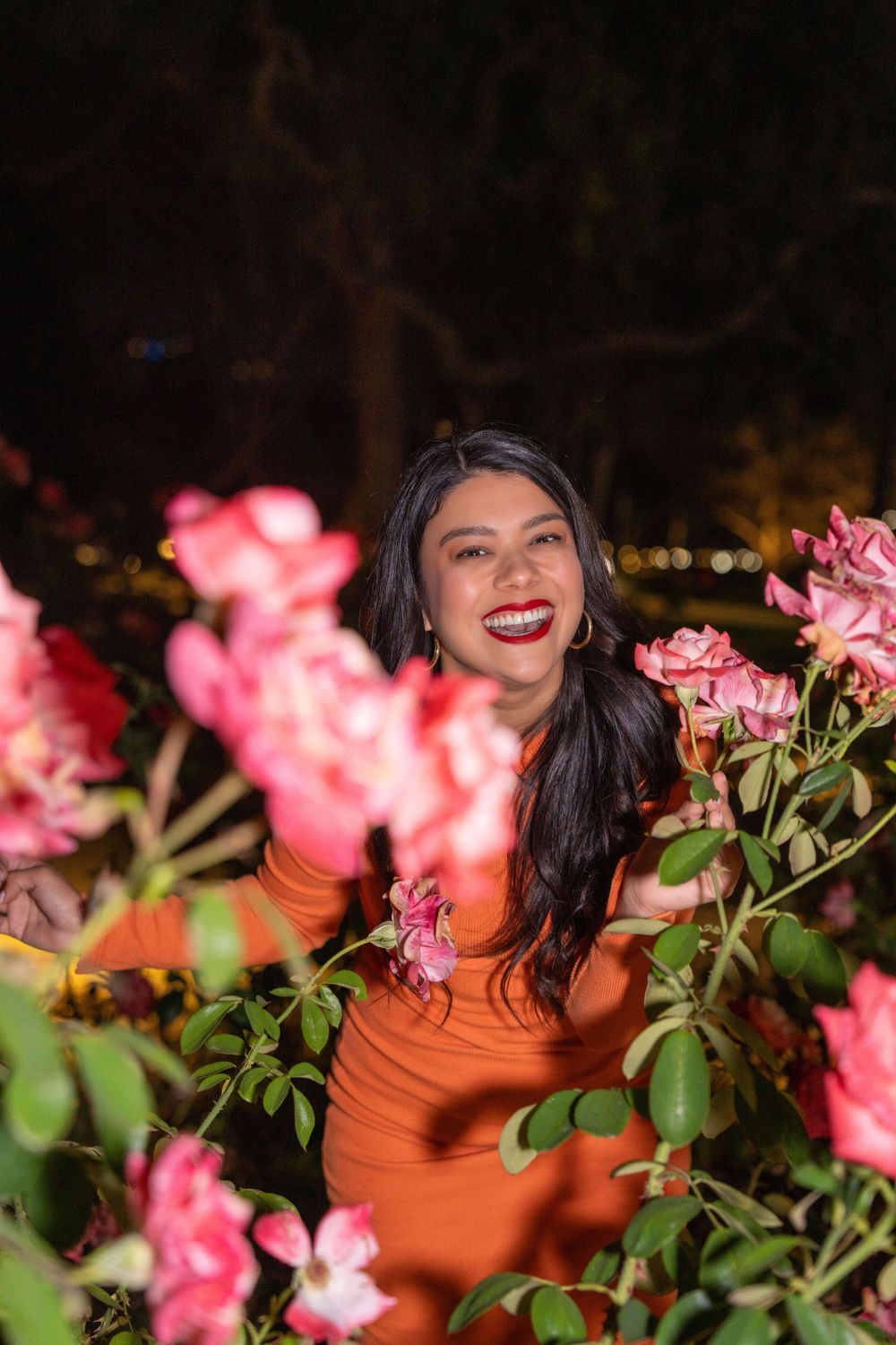 Bright smile captured through pink roses as woman poses in orange dress.