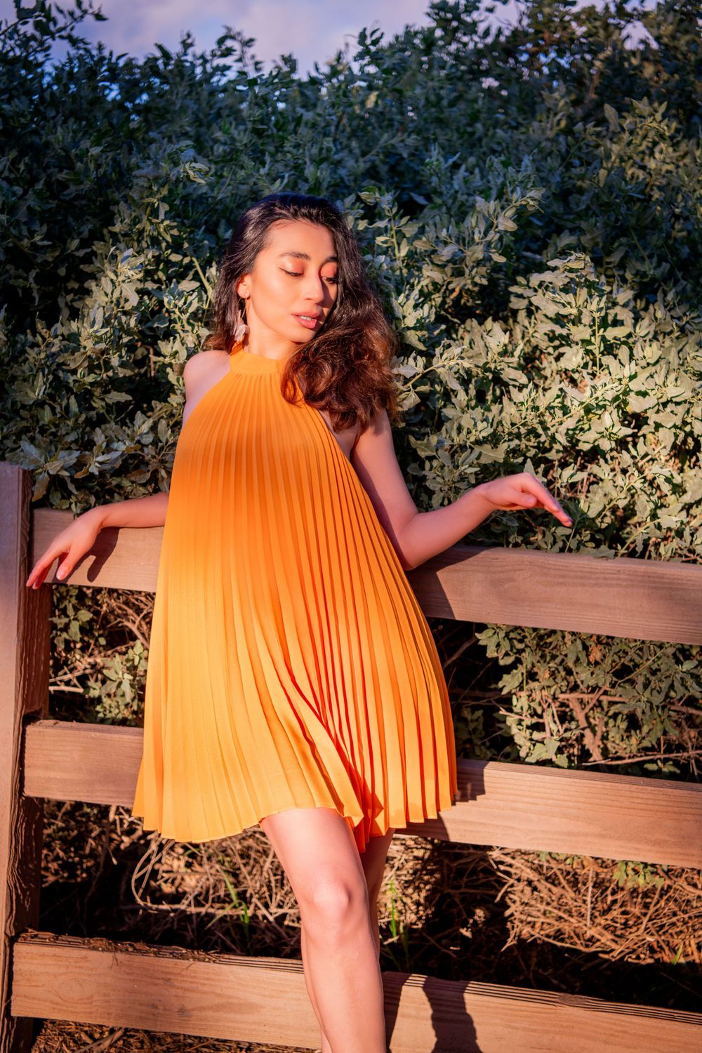 Graceful pose in orange pleated dress against wooden fence at dusk.