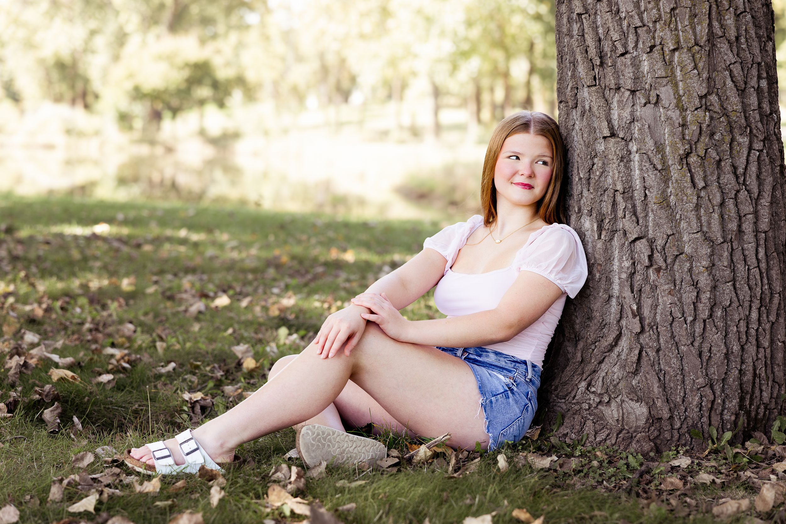 Someone sits casually against a large tree trunk on grass covered in scattered autumn leaves in natural sunlight.
