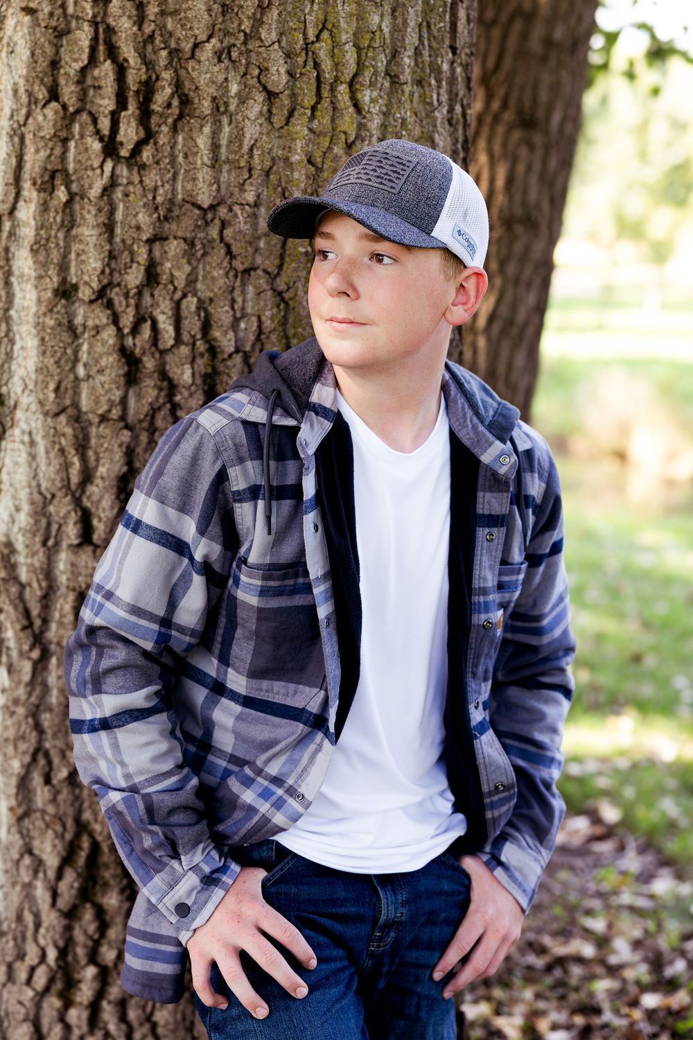 Casual outdoor portrait against a large tree trunk wearing plaid flannel, white t-shirt, baseball cap and jeans.