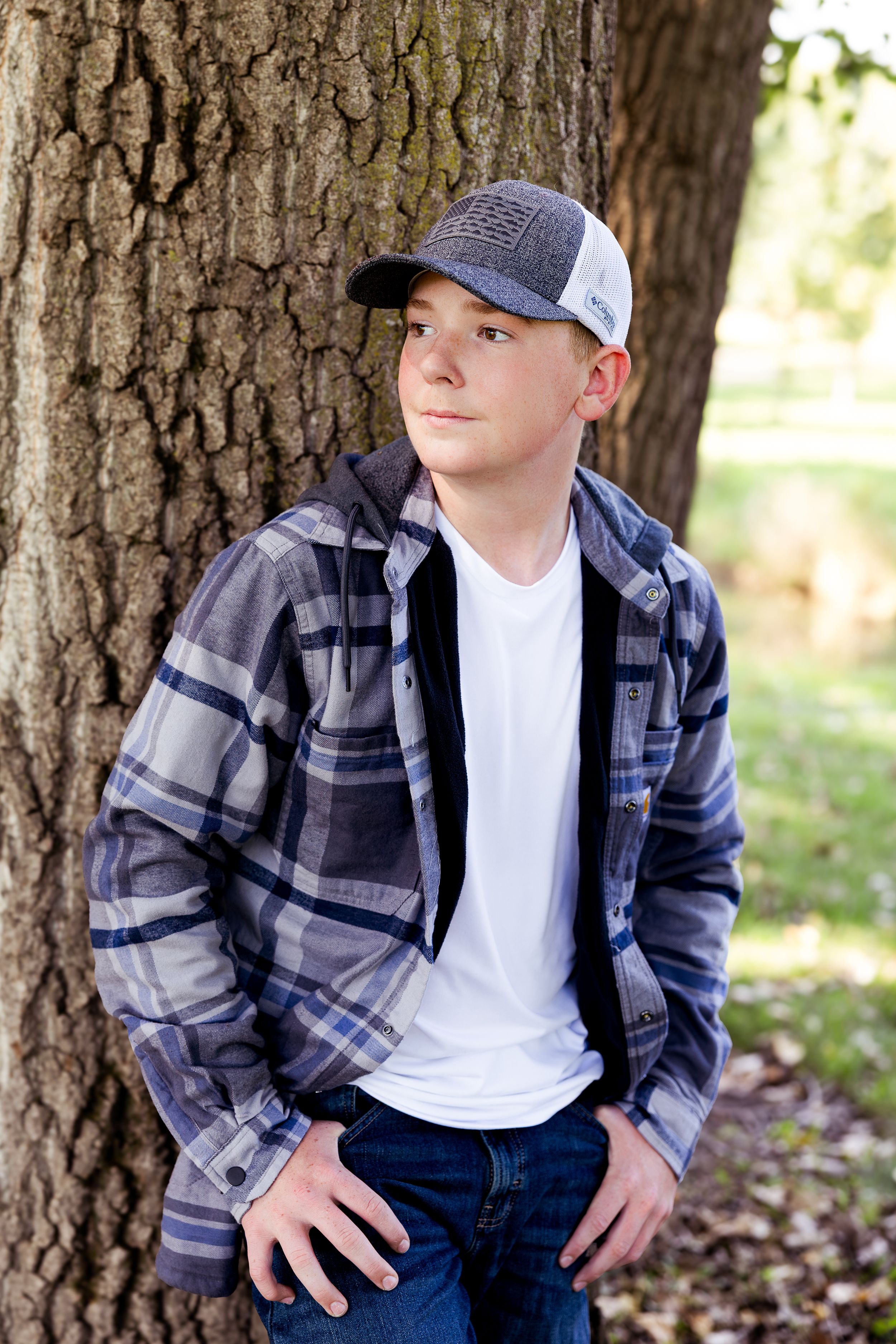 Casual outdoor portrait against a large tree trunk wearing plaid flannel, white t-shirt, baseball cap and jeans.