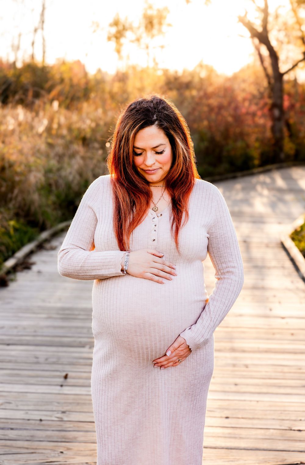 A pregnant woman in a cream sweater stands on a wooden boardwalk during golden hour.