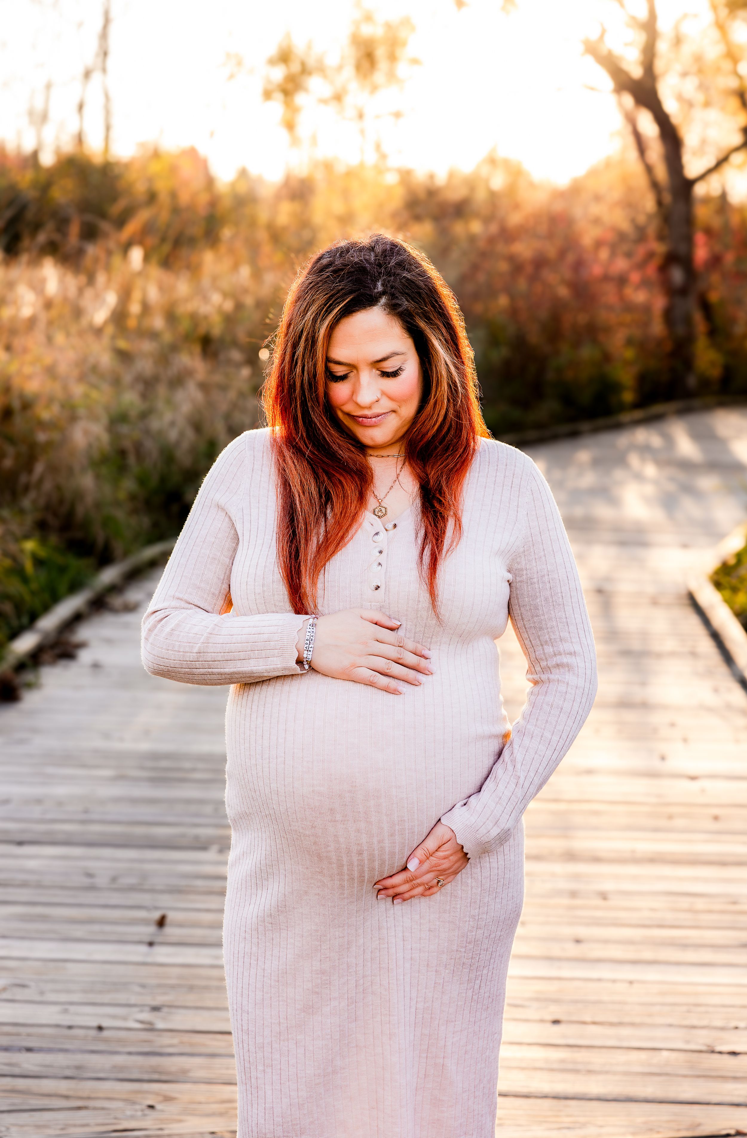 A pregnant woman in a cream sweater stands on a wooden boardwalk during golden hour.