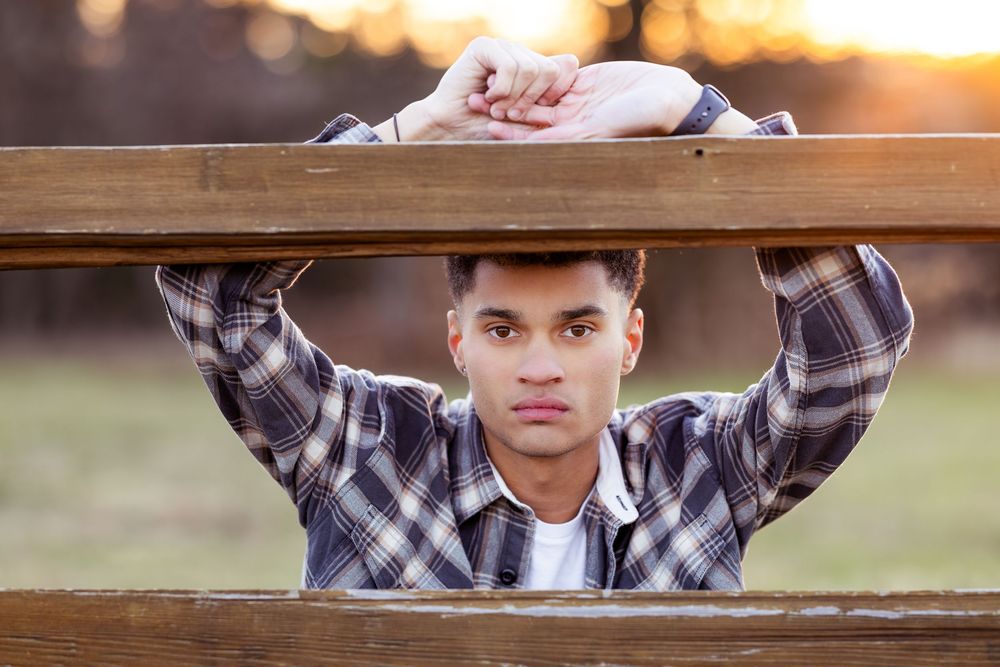 A person in a plaid shirt stands behind a wooden fence at sunset with warm golden light in the background.