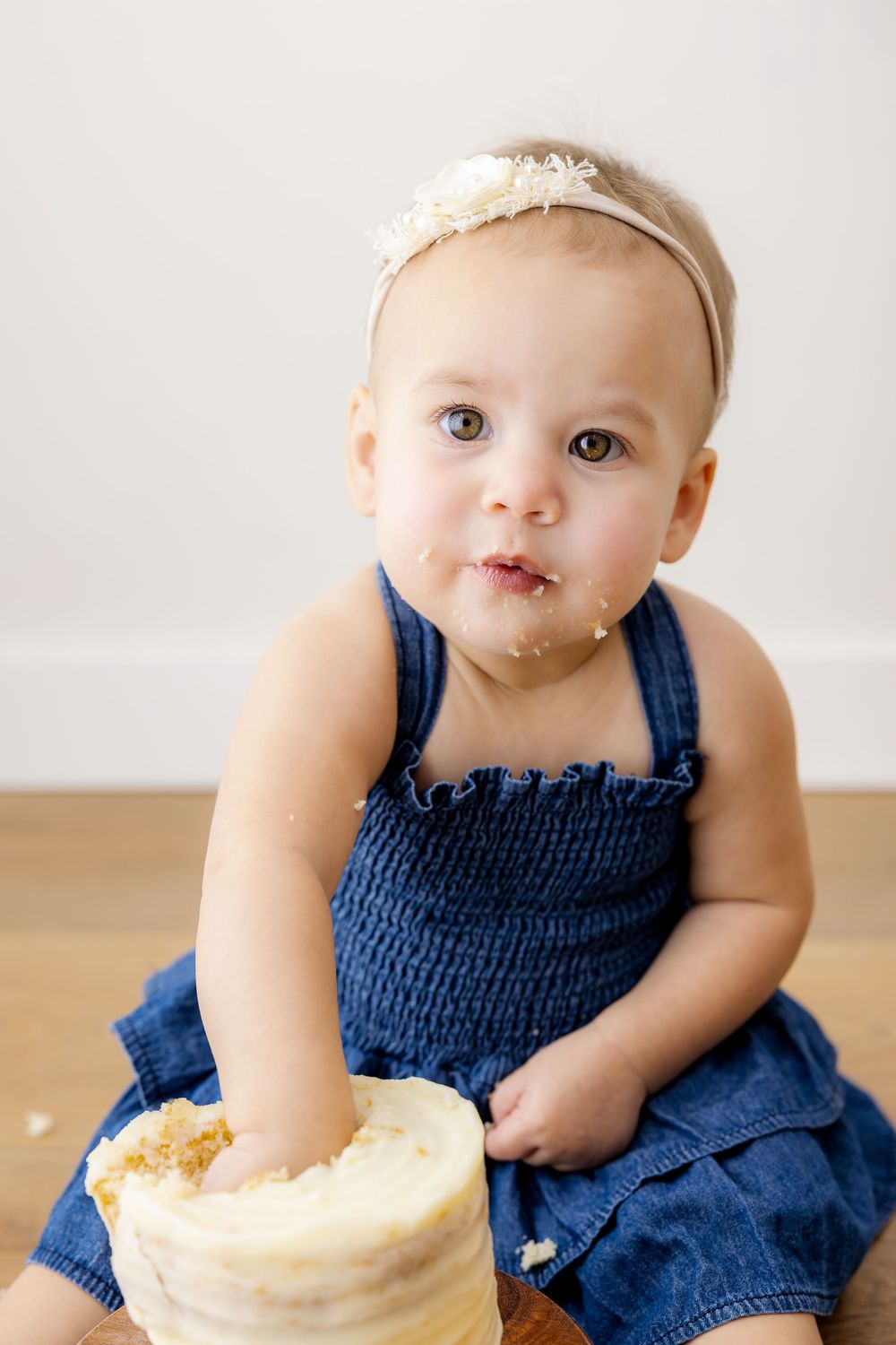 Baby in navy blue dress sits on floor with birthday cake and messy frosting smeared on their face.