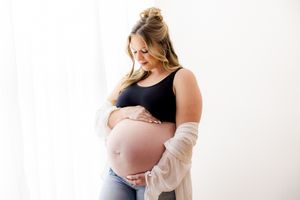 Pregnant woman in black tank top and jeans cradling her bare baby bump near a bright white window.