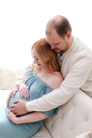 A couple in white and blue share a tender moment during a maternity photoshoot on a light colored couch.