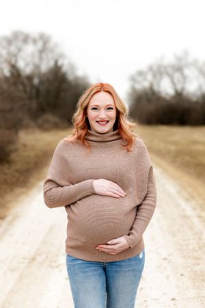 A pregnant person in a brown turtleneck sweater and jeans stands on a dirt path during a maternity photo shoot.