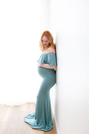 Expectant mother in flowing teal off-shoulder maternity gown poses elegantly against white backdrop.