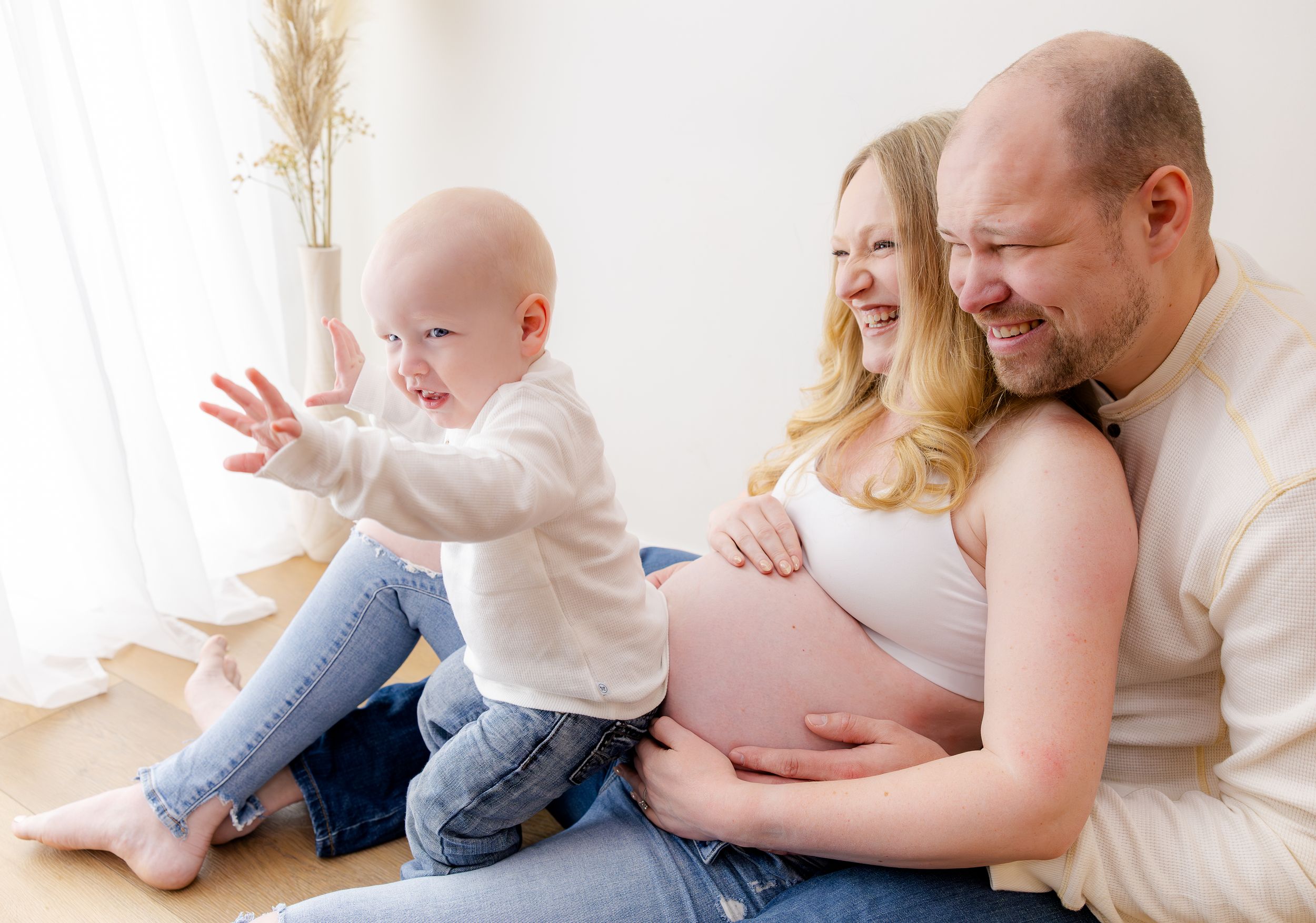 Happy pregnant family with bald husband, smiling blonde wife, and excited toddler touching baby bump in bright white room.