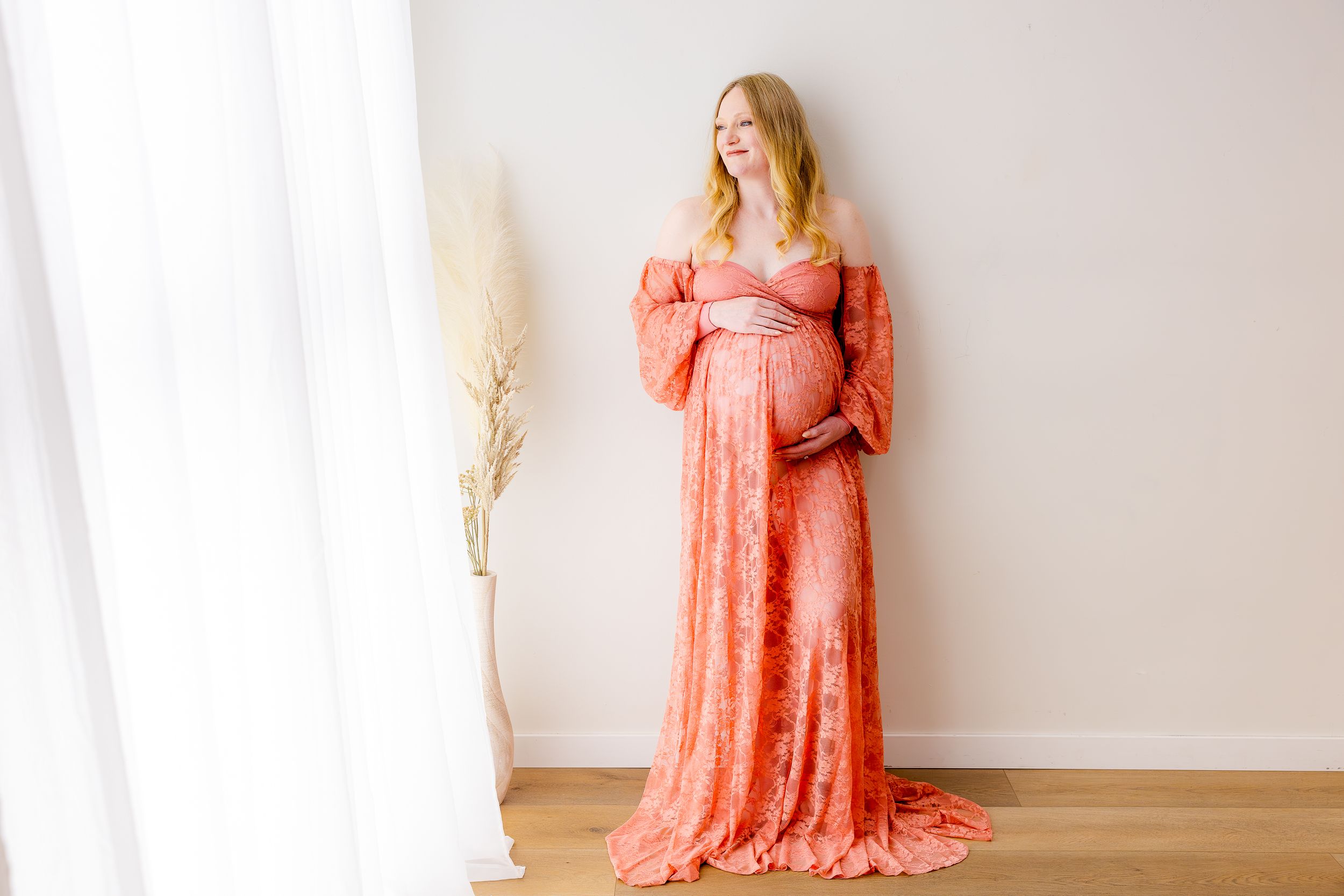 Pregnant woman in elegant coral lace maternity gown posing gracefully near sheer white curtains in a bright studio.