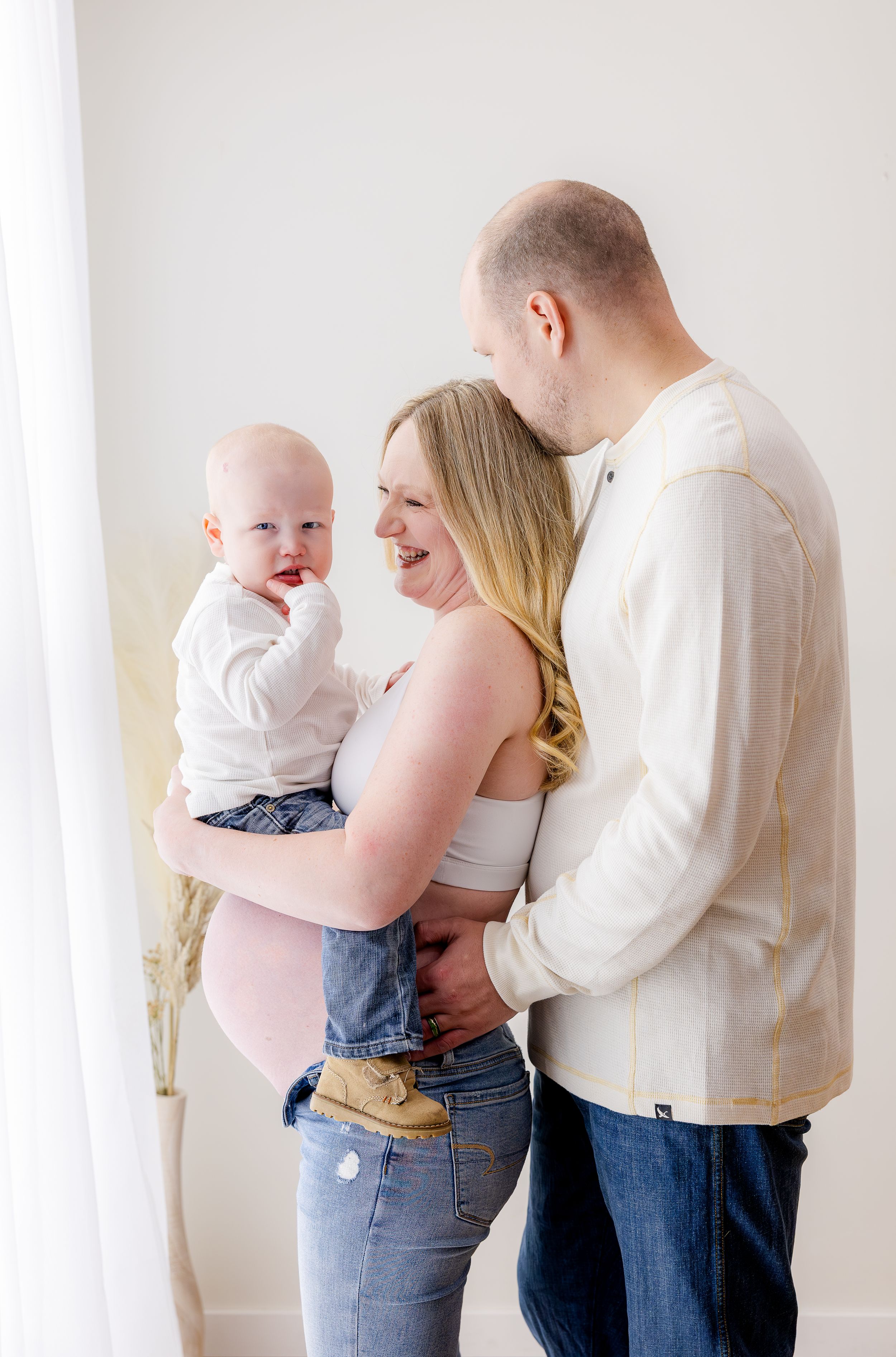 Pregnant woman holding toddler boy smiles as husband embraces her from behind in bright white studio maternity photo.