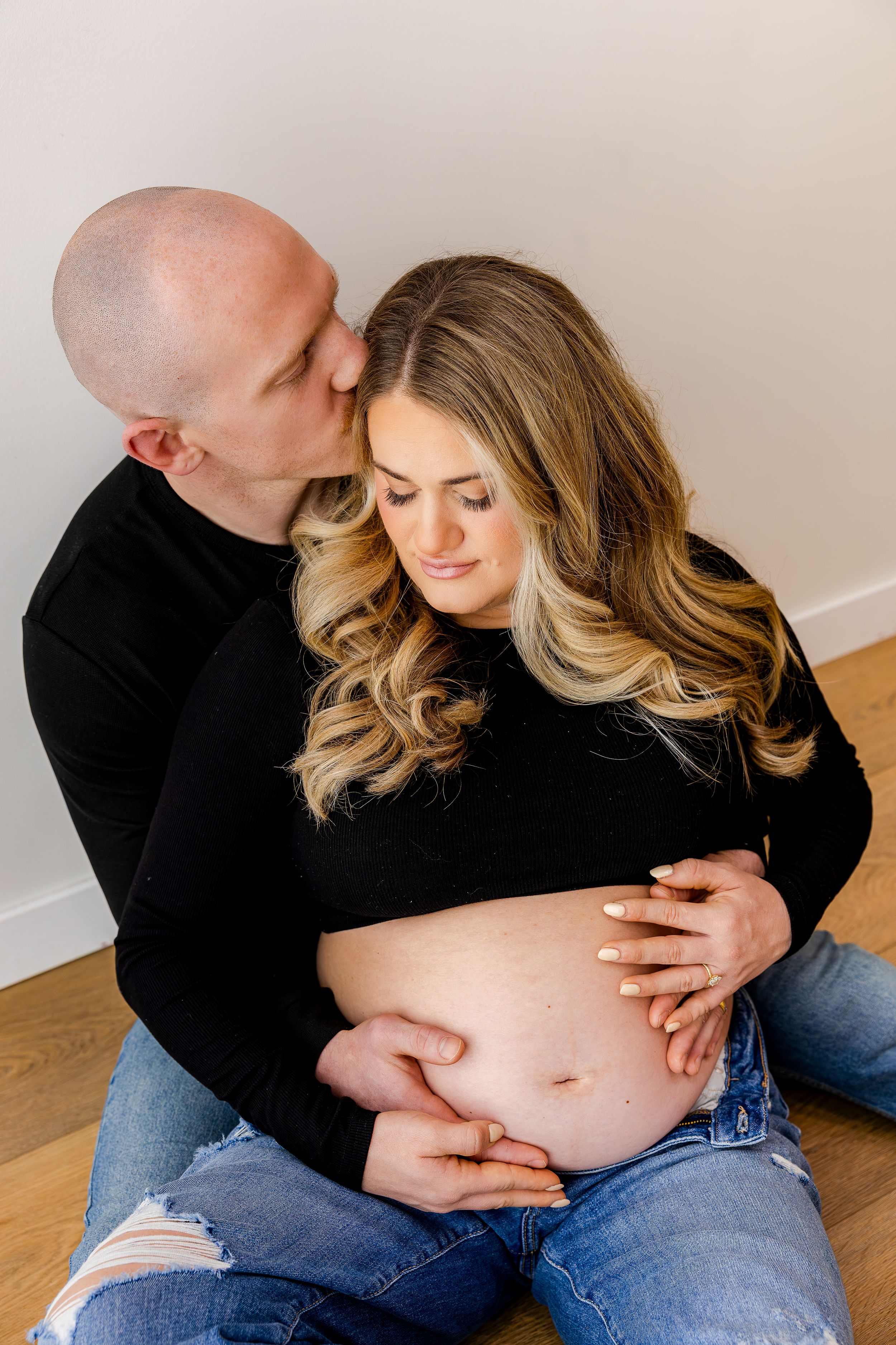 Couple sharing a tender maternity moment, man kissing pregnant woman's cheek while both cradle her bare baby bump.