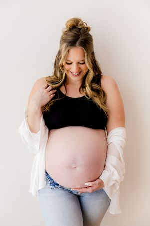 Smiling pregnant woman in black crop top, white cardigan, and jeans cradling her baby bump against a light background.