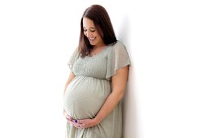 A pregnant person in a flowy light sage green dress stands against a white background, holding their belly with a gentle smile.