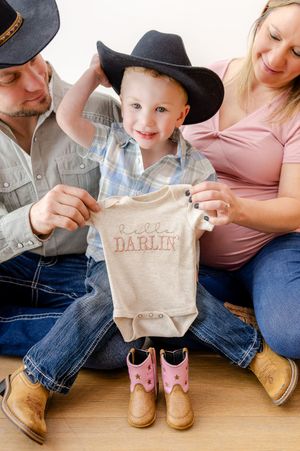 A toddler in a cowboy hat holds a Hello Darlin onesie with parents during a country-themed pregnancy announcement photo.