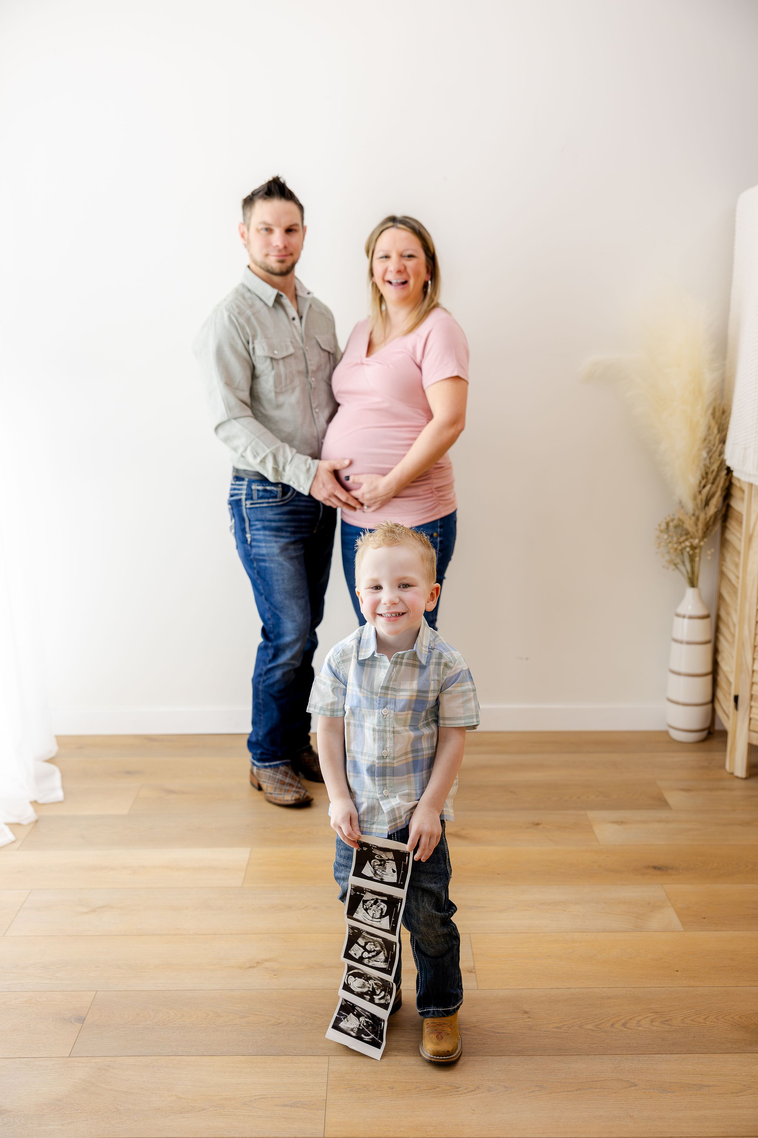 Pregnant woman and man pose with young boy holding ultrasound photos in bright modern room.