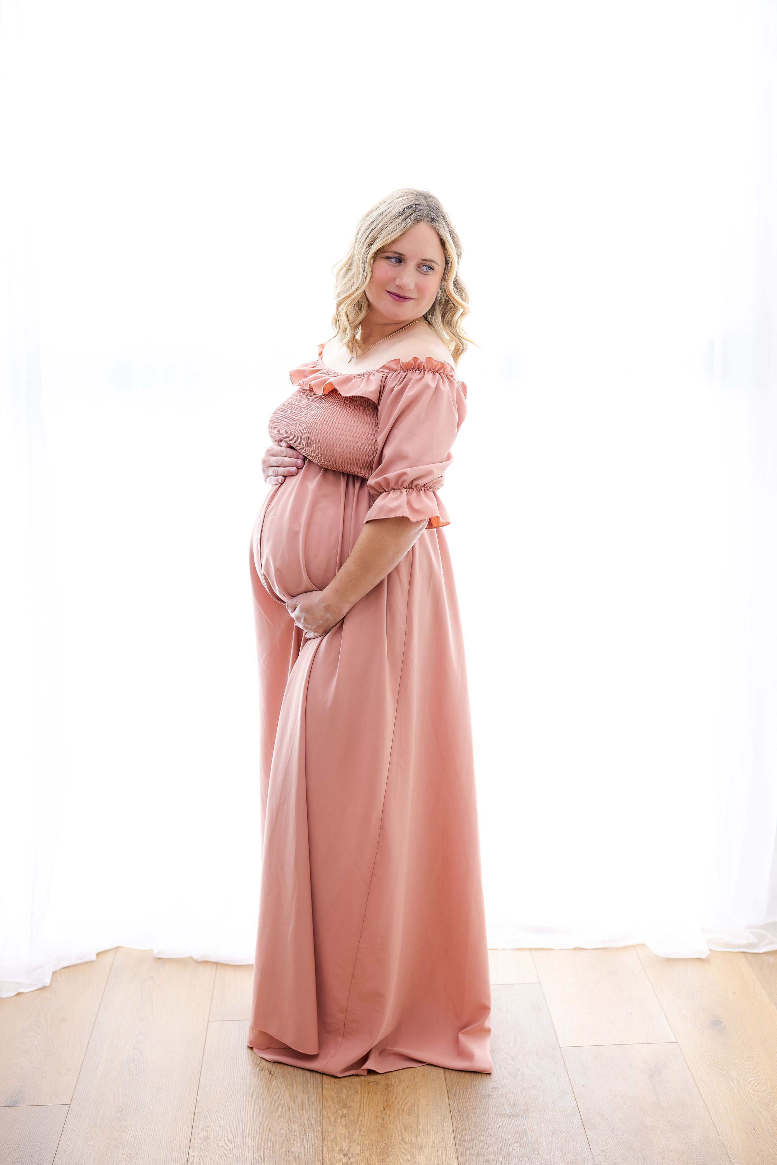 A pregnant woman poses in a blush pink off-shoulder maxi dress with ruffle details on wooden floor against white background.