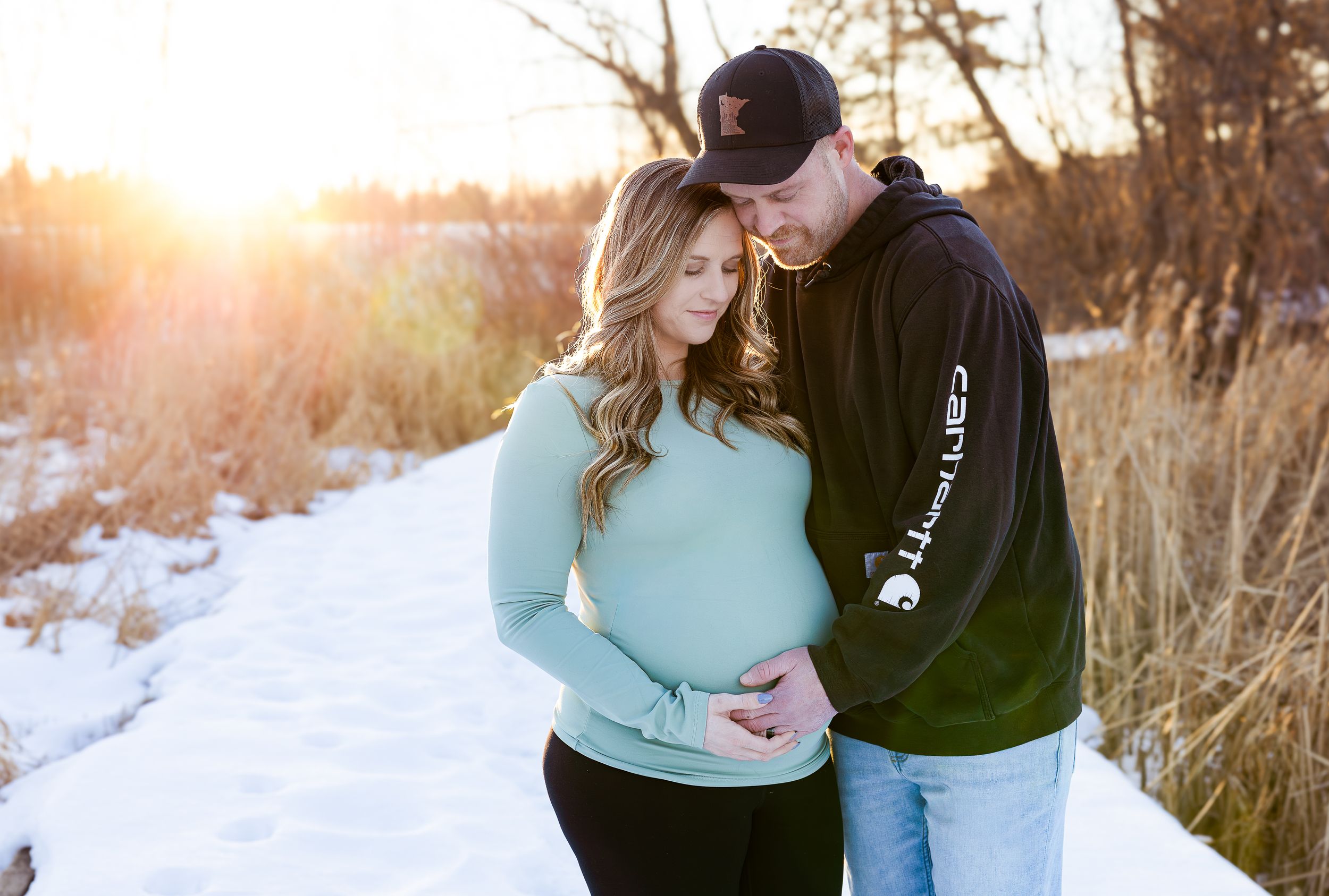 Couple sharing a tender moment in snowy winter landscape at golden hour, woman in mint sweater showing baby bump.