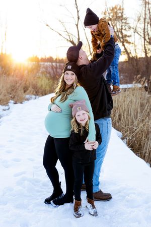 Pregnant family of four posing on snowy path at sunset, mom in teal top, dad lifting toddler, young girl smiling in front.