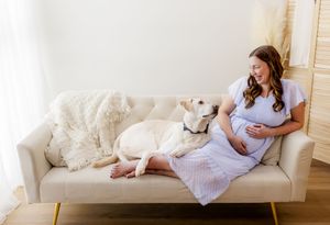 Pregnant woman in blue dress sitting on beige sofa with white Labrador dog, smiling in bright airy room.