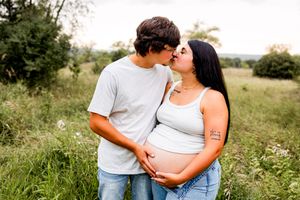 Pregnant couple sharing a kiss outdoors in a green field, man tenderly holding woman's baby bump during maternity photo session.