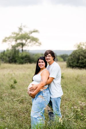 A couple poses in a grassy field, the man embracing his pregnant partner from behind, both smiling warmly outdoors.