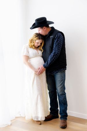 Intimate maternity portrait of couple in white dress and western wear standing against plain white wall.