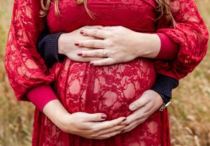 Couple tenderly cradling pregnant belly in red lace maternity dress during outdoor fall photoshoot.