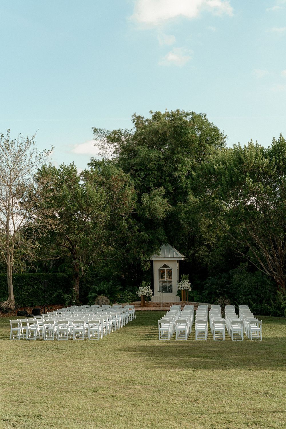 Outdoor wedding ceremony setup with white chairs arranged on a lawn facing a gazebo.