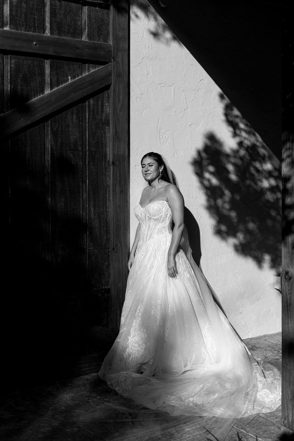 Black and white artistic portrait of a bride in a flowing white gown against a shadowed wall.