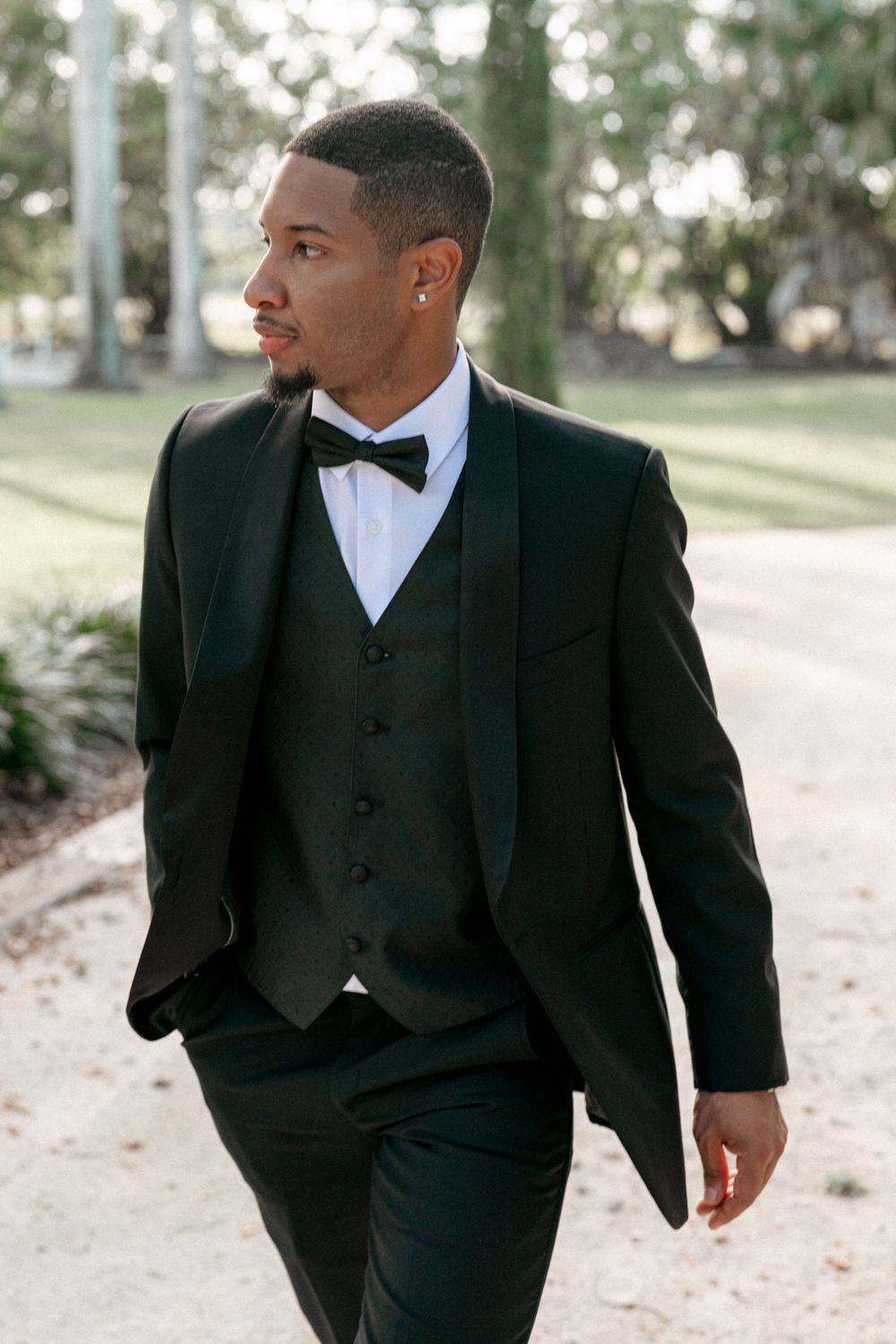 Formal black three-piece tuxedo with bow tie worn at an outdoor wedding venue.