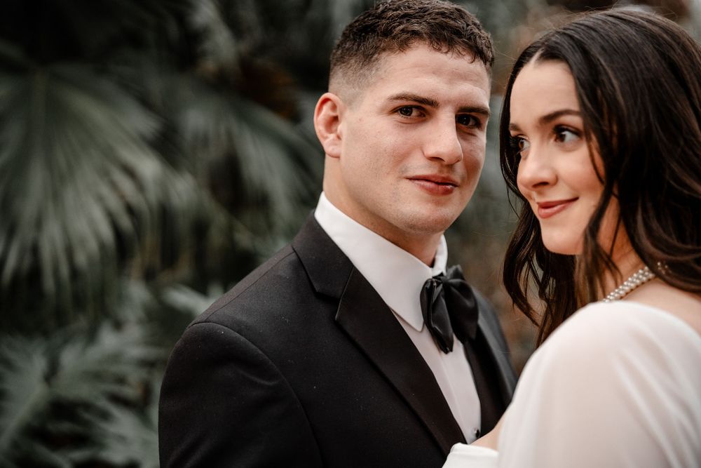 A romantic wedding portrait of a couple in formal attire standing close together with tropical foliage in the background.