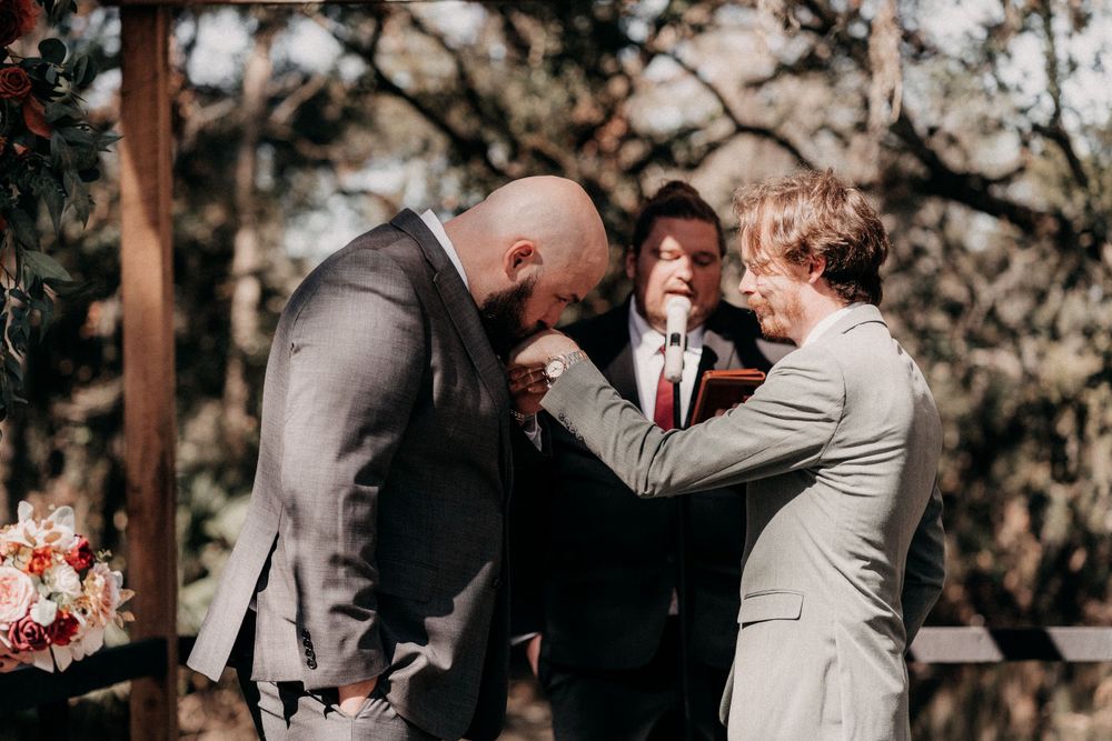 Three people in formal attire share an emotional moment during an outdoor wedding ceremony.