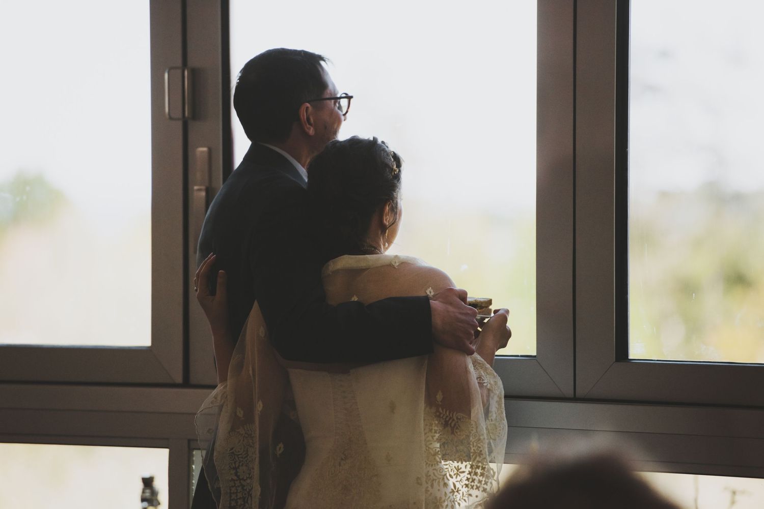 Couple embracing while looking out large windows during wedding reception.